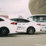 Białe samochody usług przewozowych Uber stoją zaparkowane obok siebie na otwartym parkingu. Na zdjęciu widać trzy modele: sedan Uber Comfort, sedan Uber Black oraz większy van oznaczony jako UberXL. Wszystkie pojazdy mają białe oznaczenia Ubera na drzwiach i dachach. W tle znajduje się nowoczesny budynek o zaokrąglonej, metalicznej konstrukcji oraz znak „WYJAZD”