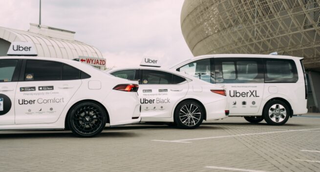 Białe samochody usług przewozowych Uber stoją zaparkowane obok siebie na otwartym parkingu. Na zdjęciu widać trzy modele: sedan Uber Comfort, sedan Uber Black oraz większy van oznaczony jako UberXL. Wszystkie pojazdy mają białe oznaczenia Ubera na drzwiach i dachach. W tle znajduje się nowoczesny budynek o zaokrąglonej, metalicznej konstrukcji oraz znak „WYJAZD”